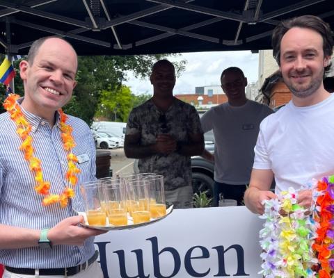 Two white men smiling in front of a stall and wearing necklaces made of flowers 