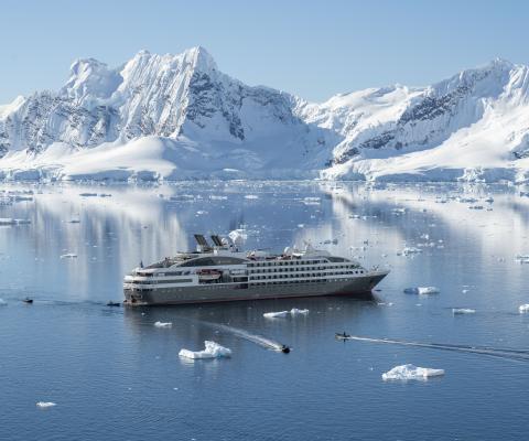 PONANT's ship L'Austral anchored among floating ice in calm blue waters, with Zodiac boats heading out and snow-covered mountains reflected in the sea behind.