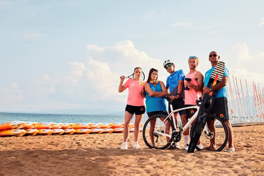 Group of people on beach holding different sporting equipment, from bikes to tennis racquets