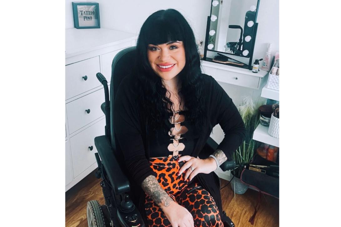 White woman with shoulder-length black hair smiling, using wheelchair, office background