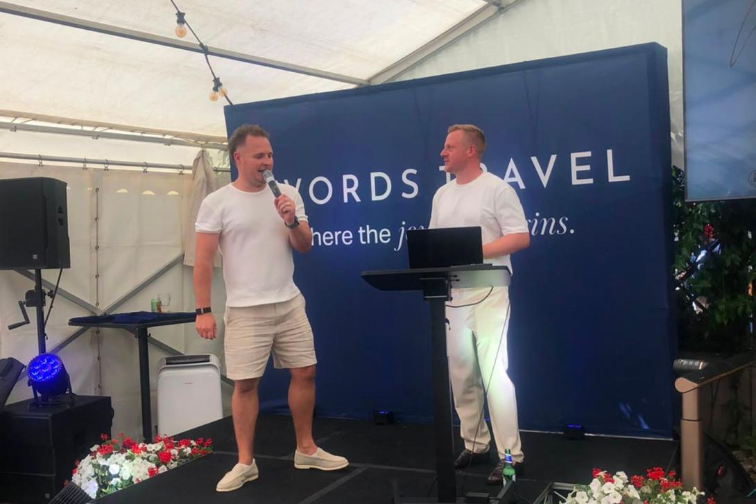 Two white men on small stage, wearing summery clothing and addressing an audience