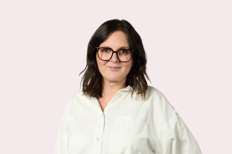Headshot of brown haired woman with glasses and white shirt, light pink background,